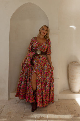 A Woman in a colorful boho dress standing in a sunlit room with an archway and vase.