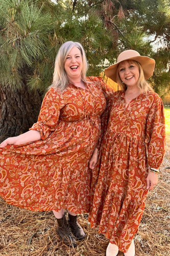 Two women in orange boho patterned dresses standing next to a tree.