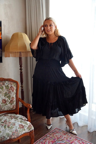 Woman in a black dress standing in a room with a lamp and chair.