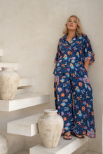 Woman wearing a blue boho floral patterned outfit standing in a minimalistic room with white shelves and vases.